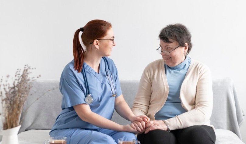 A friendly hospice nurse in blue scrubs holding the hands of an elderly woman while sitting together on a white sofa.