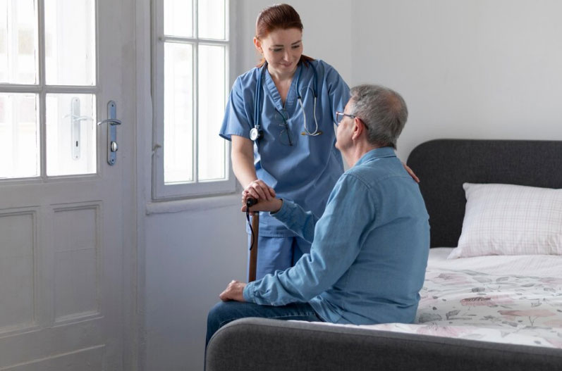 A hospice nurse in blue scrubs providing personalized care to an elderly male patient sitting on his bed in a home setting.