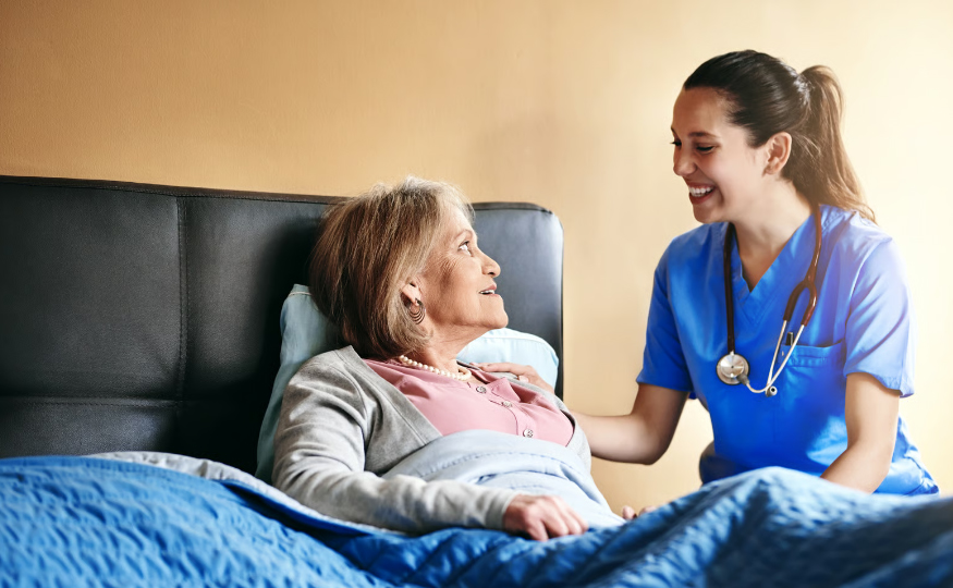 A smiling female nurse in blue scrubs with a stethoscope talking to an elderly woman resting in bed during a home healthcare visit.