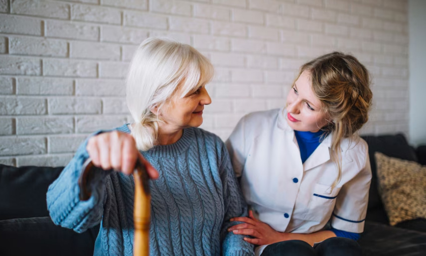 A hospice care provider offering emotional and spiritual support to an elderly woman sitting on a sofa.