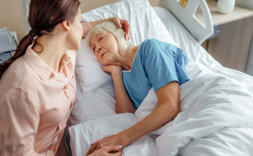 A young woman in a pink shirt leaning over an elderly woman in a hospital bed, gently touching her head and holding her hand to provide comfort during breathing changes.
