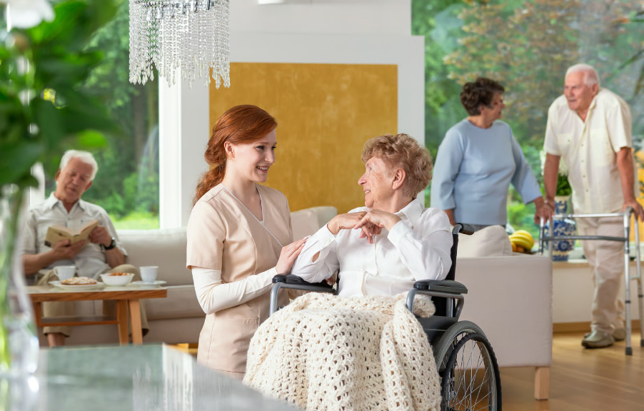 A smiling young redhead female caregiver in a beige uniform talks with an elderly woman in a wheelchair under a blanket, while other senior residents relax, read, and walk with a walker in a bright nursing home lounge.