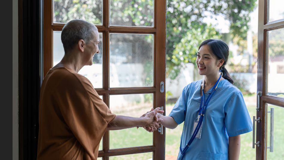 A smiling young Asian female nurse in blue scrubs with a stethoscope around her neck shakes hands at a glass doorway with a smiling cancer patient or senior woman who has short cropped hair.