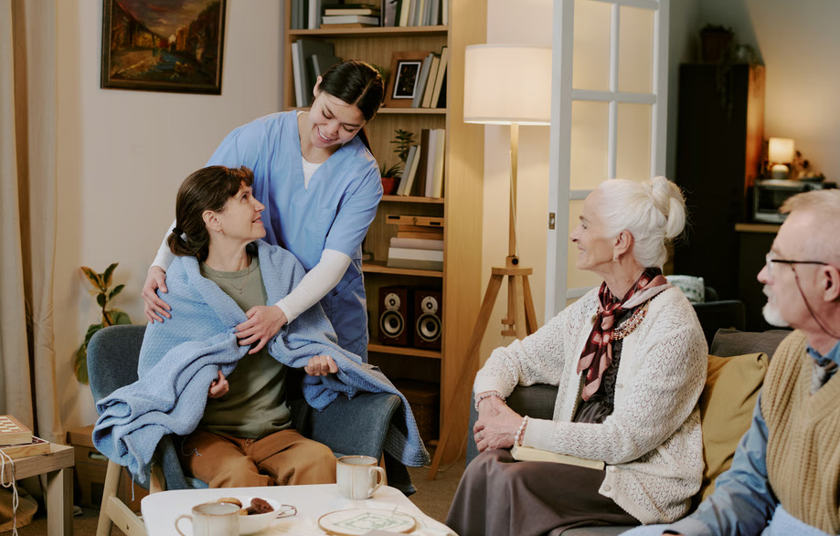 A smiling hospice nurse in blue scrubs wraps a comforting blue blanket around a female caregiver while an elderly couple sits nearby in a cozy living room.