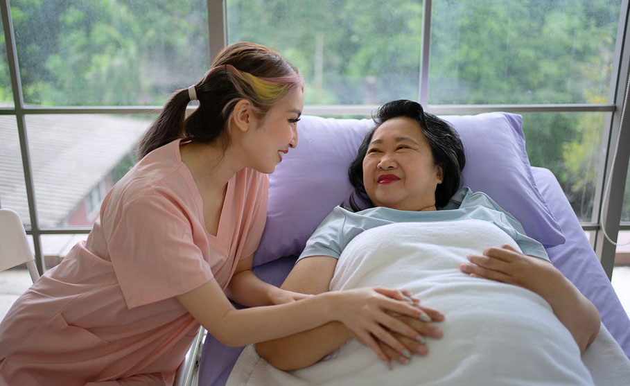 A young female nurse in pink scrubs smiling warmly while holding the hand of an elderly Asian woman resting in a hospital bed, representing early palliative care support provided by Homage Hospice Plus in the Dallas-Fort Worth area.