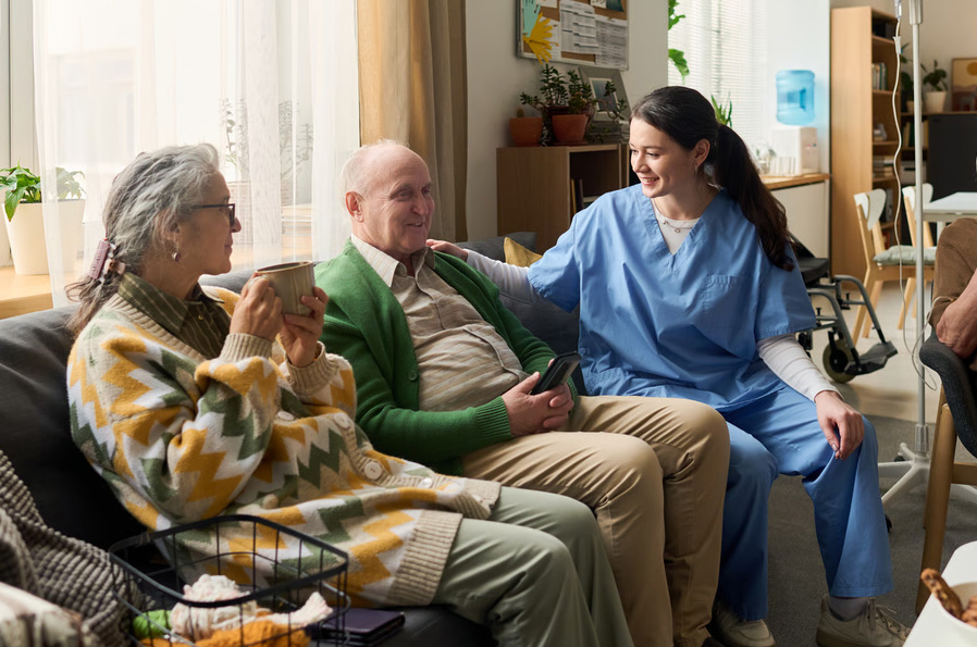A smiling female nurse in blue scrubs sits and speaks with an elderly man and woman on a couch during a supportive home care visit.