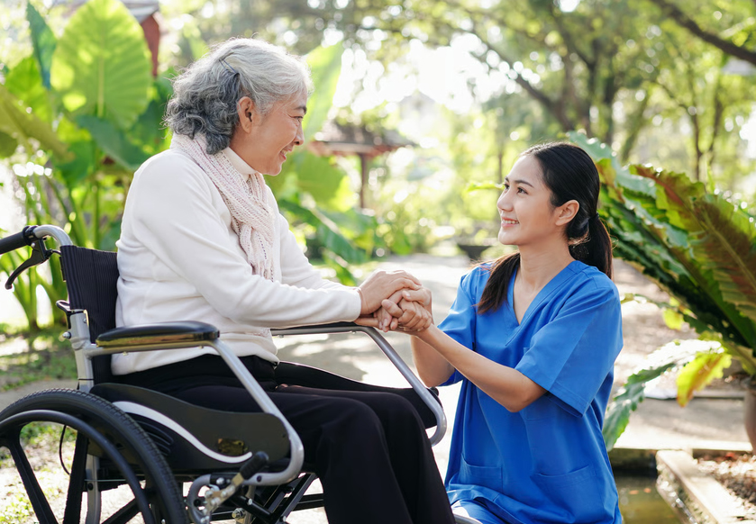 A Homage Hospice Plus caregiver in blue scrubs holding hands with an elderly senior woman sitting in a wheelchair outdoors.