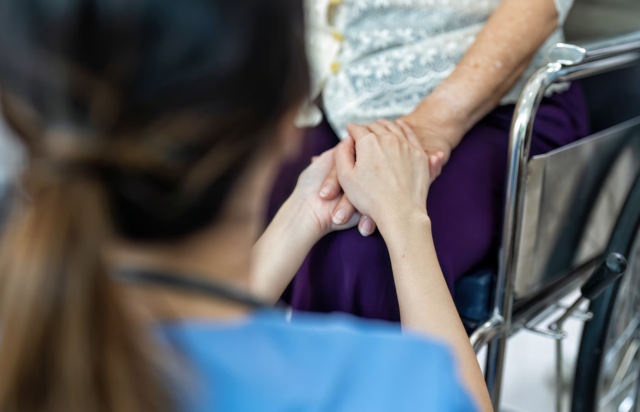 A Homage Hospice Plus caregiver holding the hands of an elderly patient in a wheelchair to offer emotional comfort and grief support.