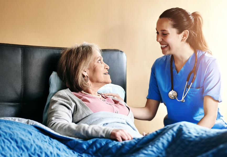 Smiling female nurse in blue scrubs talking to a senior female patient lying in bed, illustrating supportive palliative and hospice care.
