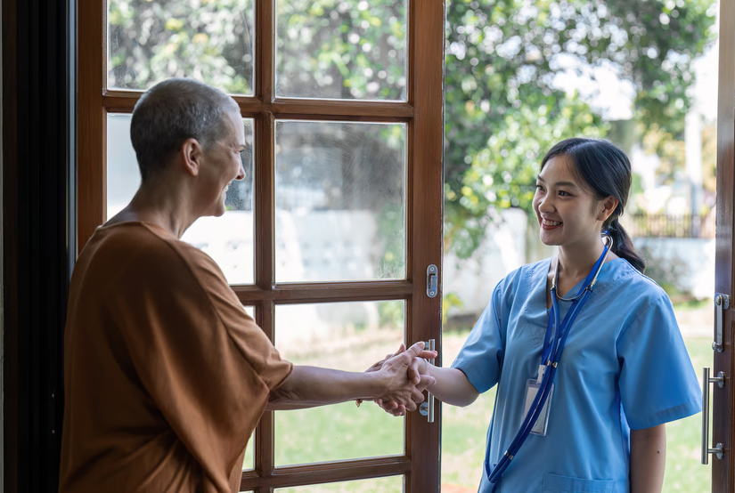A home health nurse in blue scrubs wearing a stethoscope shaking hands with a senior patient at the front doorway during a home care consultation.